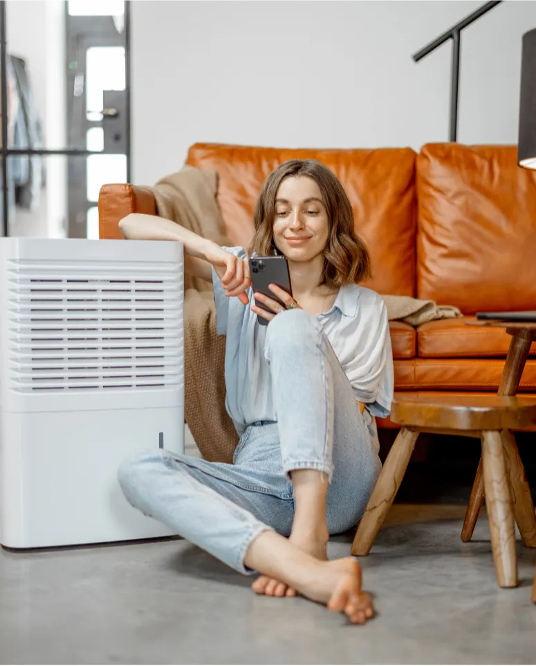 air cleaners near me: Woman sitting on the floor by a dehumidifier, smiling while using her smartphone in a cozy living room.
