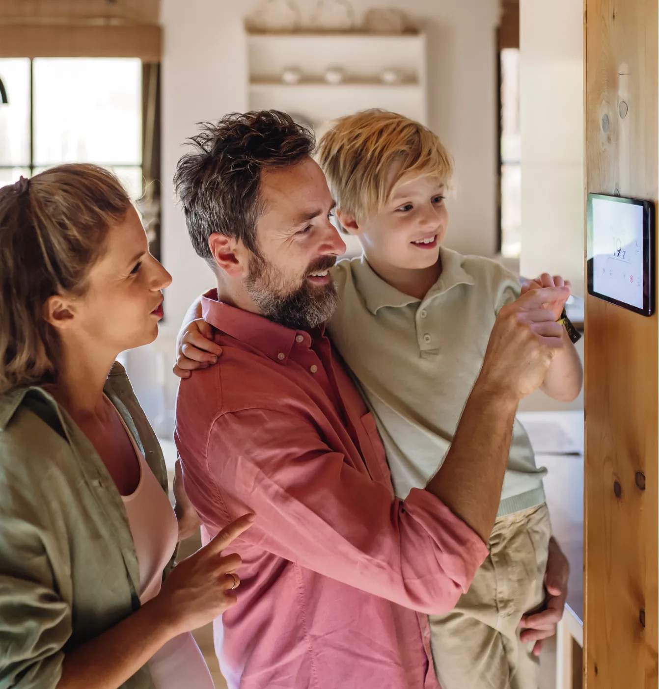 A family of three uses a smart home touchscreen panel together in a bright, modern kitchen.