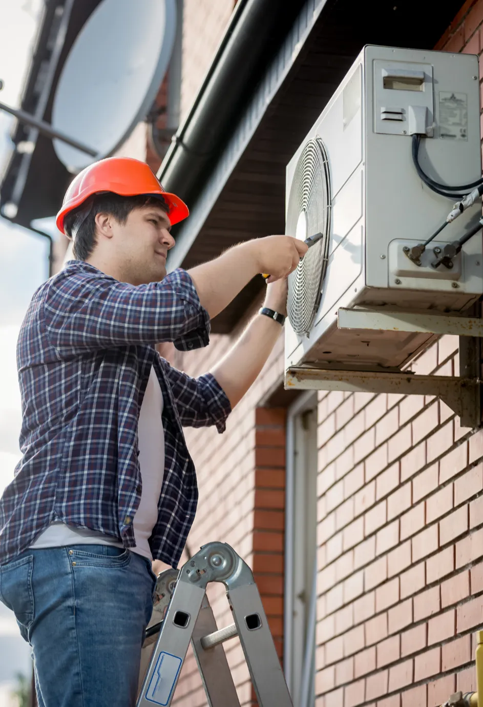 Man in a hard hat repairs an outdoor air conditioning unit while standing on a ladder by a brick wall.