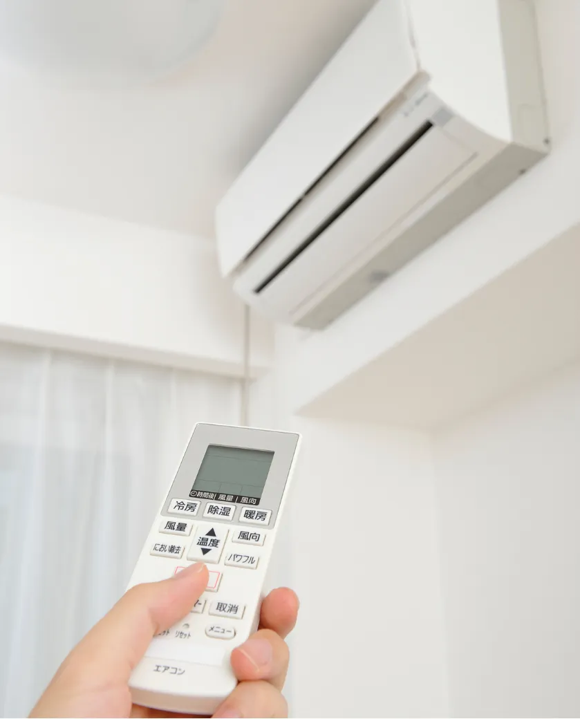 Hand holding a remote control pointed at a wall-mounted air conditioner unit in a bright room.