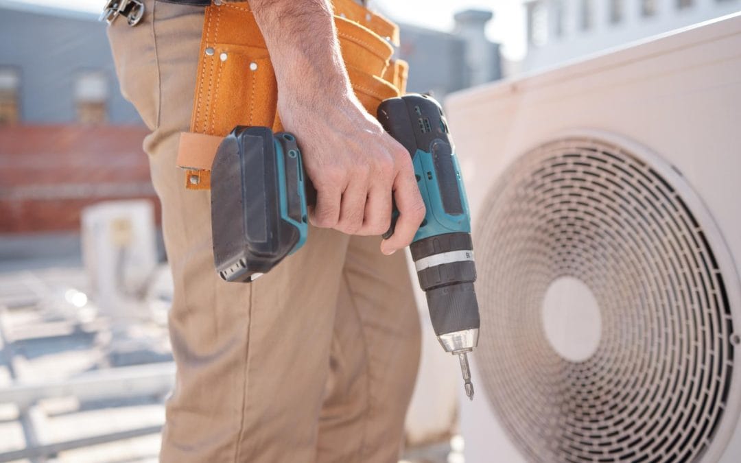 Person holding a cordless drill next to an outdoor air conditioning unit, wearing a tool belt.