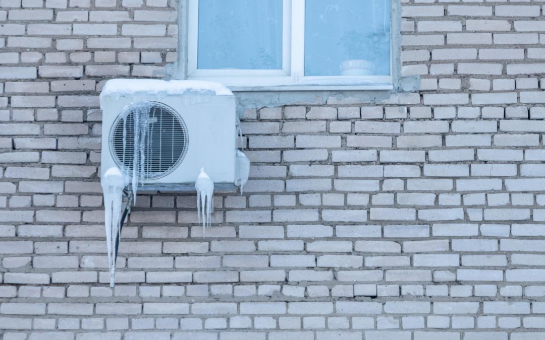 Air conditioner unit covered in ice and icicles mounted on a brick wall beneath a window.